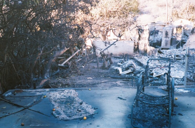 The charred remains of the Palisades Fire. A charred lawn chair sits on concrete. Behind it are ashes and burned walls.