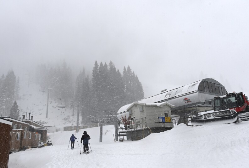 Two people covered in dark colored jackets are on skis next to a ski lift and a small structure on their left. There is white snow on the ground. Tall trees in the background. A few brown structures are to the right of the skiers.