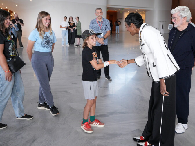 A boy wearing a Star Wars cap shakes hands with a woman in a white jacket as George Lucas and others watch nearby.