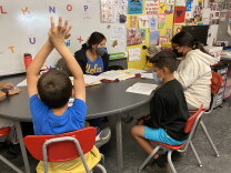 Three students seated around a semi-circular table look down at sheets of paper as their teacher, in a blue UCLA Bruins t-shirt, looks on. One student has his hands raised in a stretch.