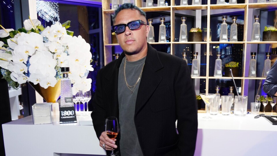 A Brown man wearing a black jacket and pants holding a beverage poses for the camera in front of a well-lit bar.