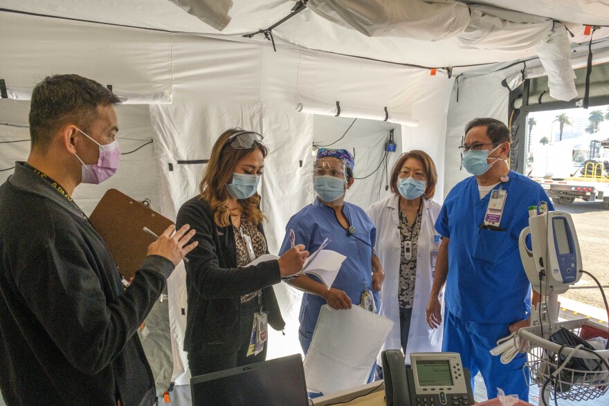 Five masked healthcare workers, some wearing white lab coats or blue scrubs, stand underneath a tent set up as a mobile ICU clinic for COVID-19 patients.