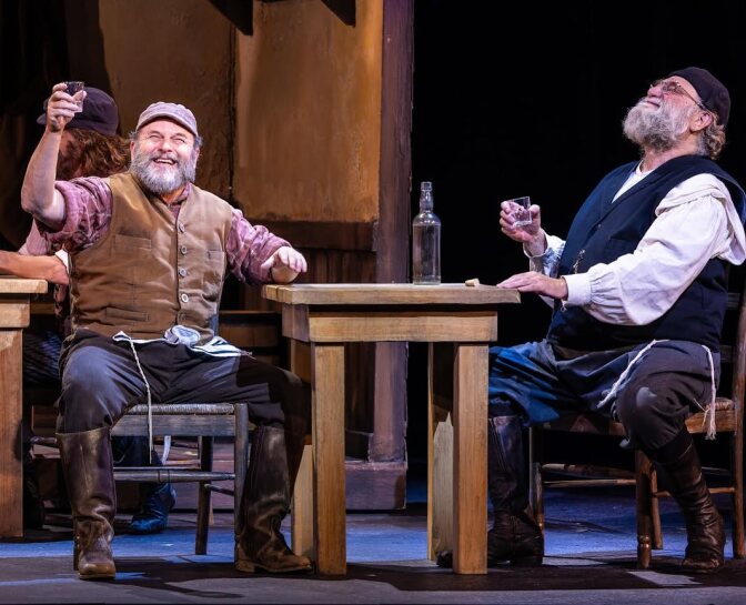 Jason Alexander and another actor sit on stage in costume, performing the musical "Fiddler on the Roof." Alexander is toasting and smiling.  