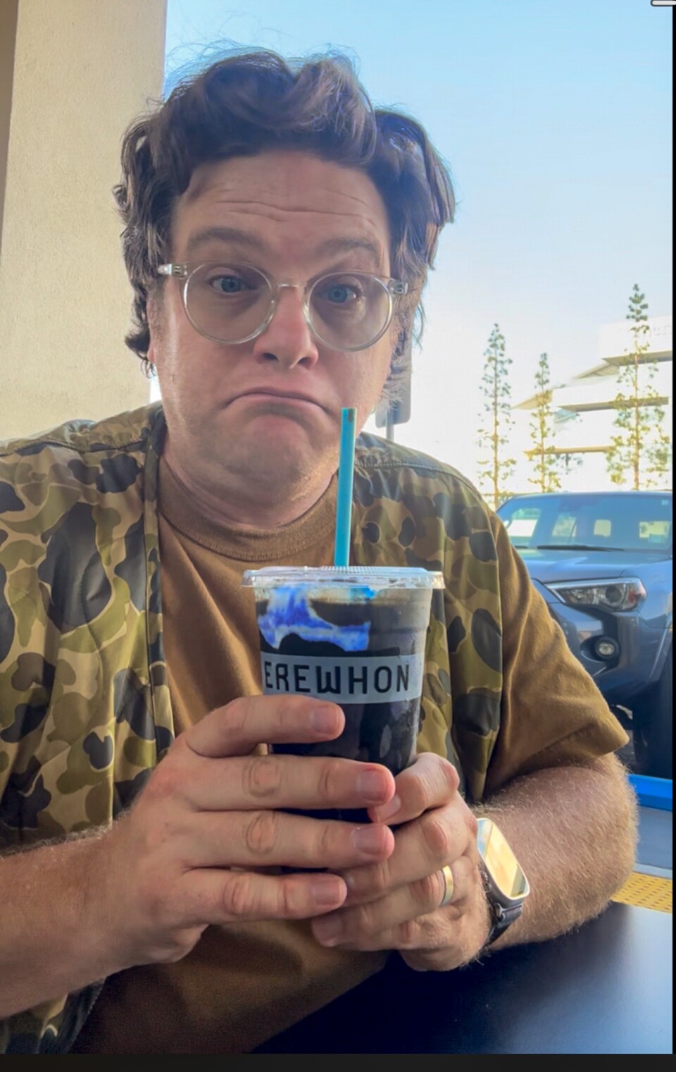 A man with a light skin tone, wearing glasses and a camo vest over a caramel-colored t-shirt, holds up a moody black-and-blue Erewhon smoothie with a mildly skeptical expression.