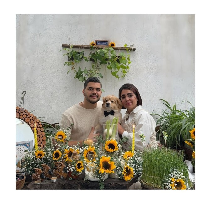 A couple sits at a table surrounded with sunflowers between a dog. 