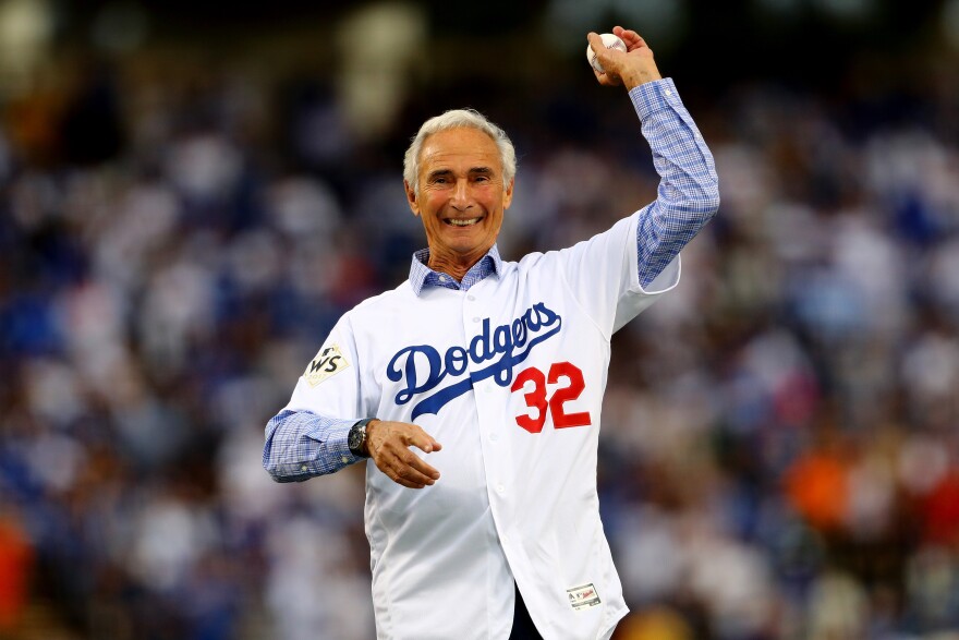 Retired pitcher Sandy Koufax, wearing a jersey that says 'Dodgers' in blue script with the number 32 below it in red over a blue dress shirt, holds a baseball in his left hand with his arm raised above his head as he prepares to deliver a pitch.