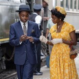 Jalyn Hall stands mid-stride on the left in a navy blue suit. He is looking down at his slightly clasped hands. His left hand has a prominent silver ring on the middle finger. On Hall's right is Danielle Deadwyler in a yellow dress with white florals. She wears a matching hat and white wrist gloves. Her right hand rests slightly below her chest as her left hand motions towards Hall's hand as if they are passing something between them.  She is also looking down at Hall's hands seemingly holding something of interest. Behind them to the left is a train car with people coming out of its doors. There is a conductor in the back right indicating that they are on at a train platform, possibly just arriving. 