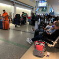 People push luggage carts and roller suitcases while others sit on chairs looking at their phone with their luggage beside them. A man wearing a dark t-shirt carries a child wearing a green t-shirt. 