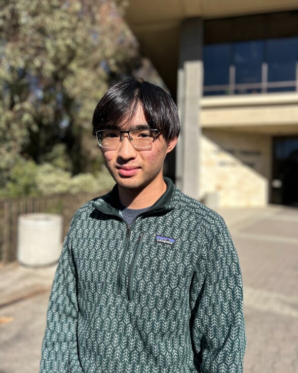 An Asian man with medium-light skin tone wearing a patterned dark green Patagonia sweater and glasses stands outside near a building. 