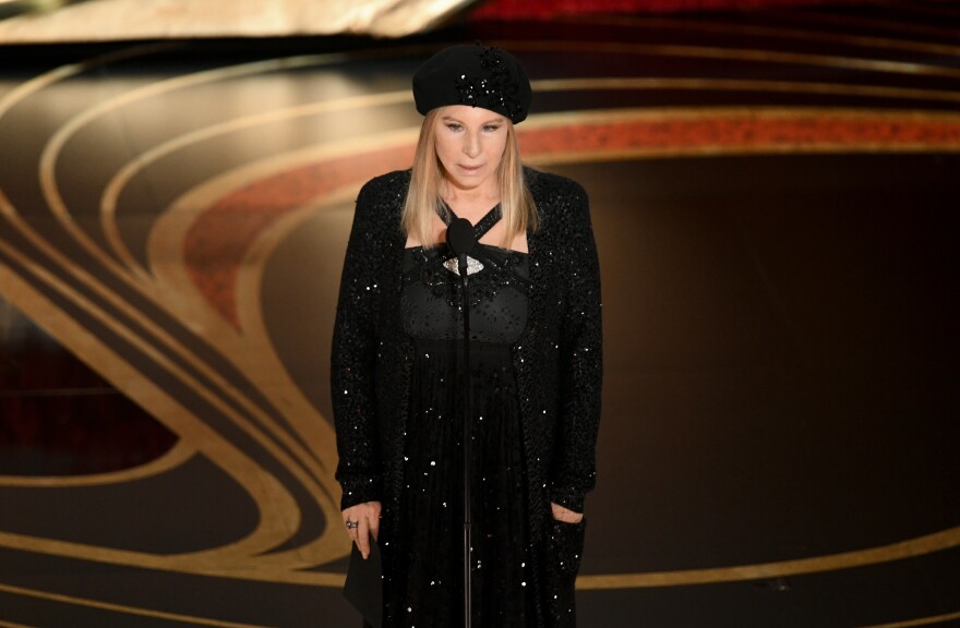 Barbra Streisand is shown onstage speaking into a microphone. She wears a long-sleeved black sequined gown and matching beret-style hat.