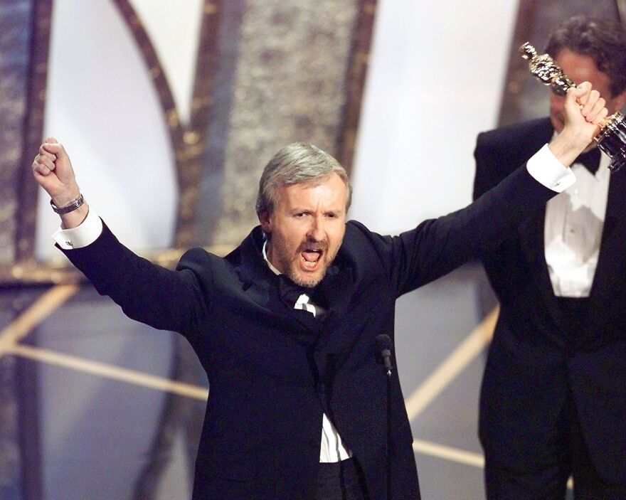 Director James Cameron has his arms raised in triumph as he holds an Oscar statue in his left hand.