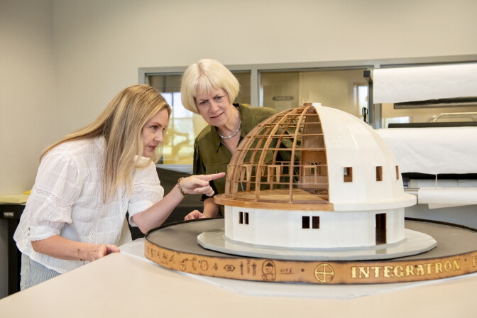 Two blonde women peer into an architectural model of a domed building. 