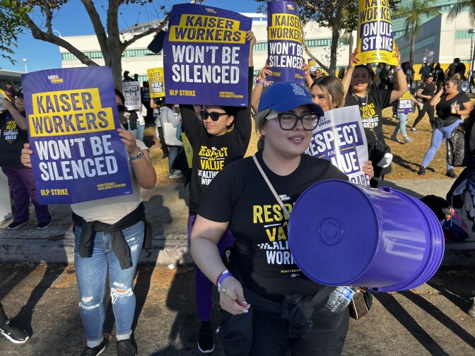 A crowd of people picket outside a hospital. They're wearing black union shirts while holding signs that read "Kaiser workers won't be silenced."