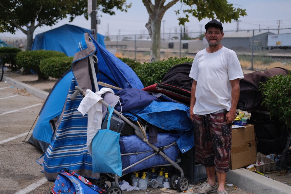 A man with light-tone skin wears short pants with the U.S. flag on them. He's standing next to a makeshift car with belongings covered in tarps.