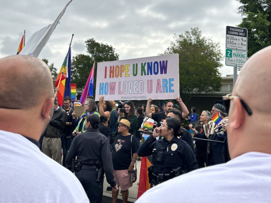 A group of LGBTQ+ supporters stand behind a police line outside the school holding up a sign that says "I hope you know how loved you are" in rainbow and trans pride flag colors.