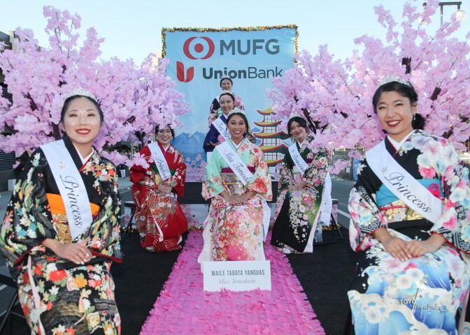 The Nisei Week queen sits on a pink carpet while her court of six flanks her. They are all wearing sashes and wear kimonos, 