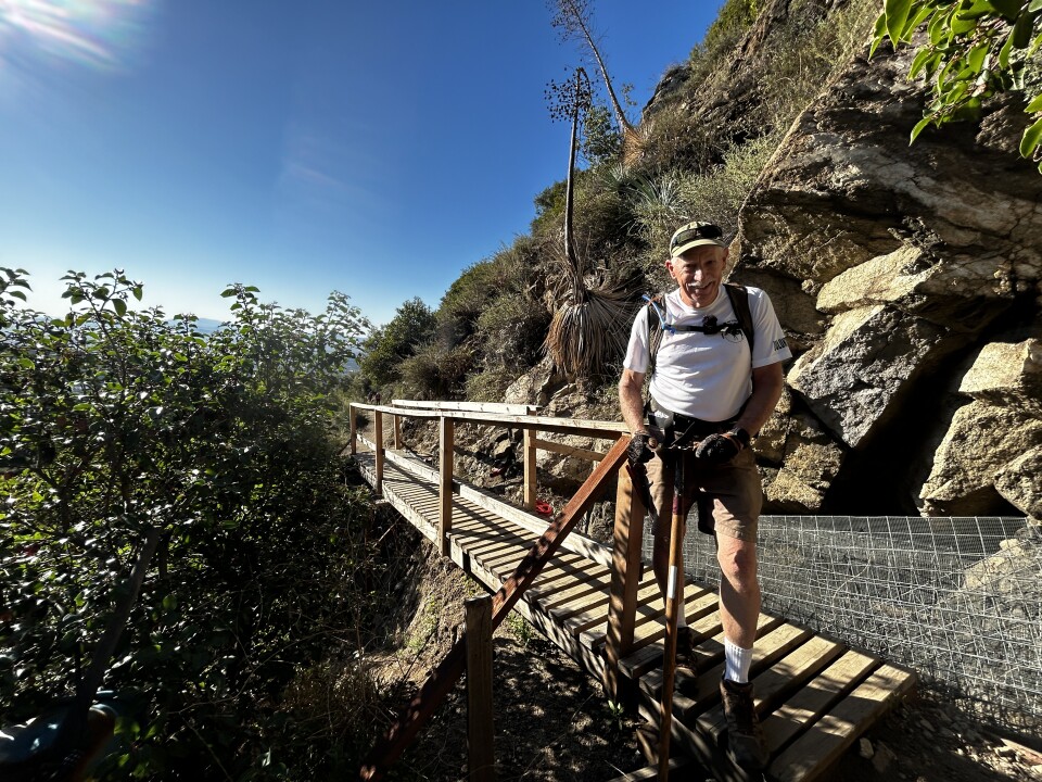 An older white man wearing a tan baseball cap and white t-shirt and tan shorts stands at the end of a new wood bridge smiling on a sunny day. In the background are large boulders, greenery and yucca plants. 