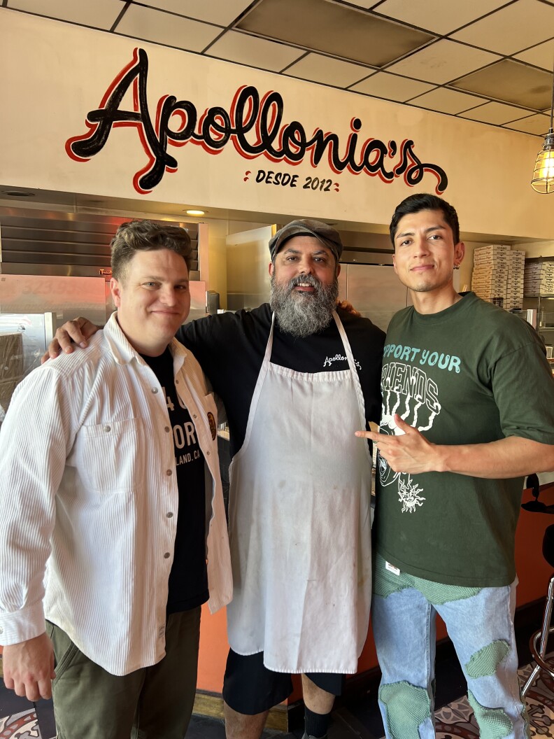 Three men stand together. The man in the center has long white and grey hair and is wearing a jaunty cap with a black T-shirt over a white apron. He has both of his arms around the men on each opposite side. The man on the far left has light skin and dark blonde hair and is wearing a white jacket over a black T-shirt. The man on the right wears a green T-shirt with a large logo in the center.