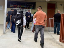 Adults and children wearing backpacks are seen from behind, walking toward the entrance of an elementary school. 
