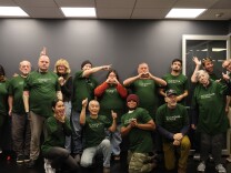 A group of more than a dozen people wearing green 'fountain house' t-shirts pose for a photo. Many are smiling and some make heart and thumbs up gestures with their hands. 