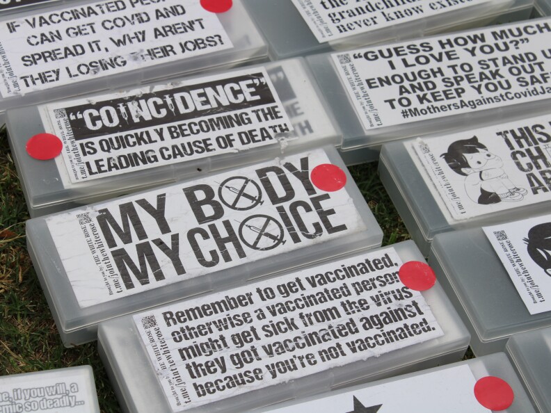 Multiple slogans and signs lay on grass reading, "coincidence is quickly becoming the leading cause of death," "my body my choice," and "remember to get vaccinated otherwise a vaccinated person might get sick from the virus they got vaccinated against because you're not vaccinated."