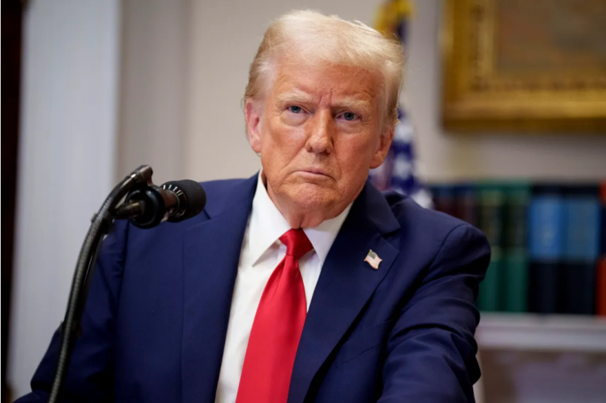 President Trump, wearing a blue suit jacket and red tie sits in front of a microphone.