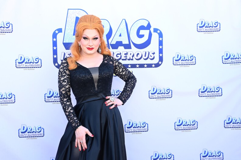Jinkx Monsoon on a red carpet poses for the camera, wearing a black dress with lacy long sleeves.