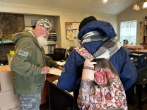 A man in an office with a full, white beard wearing an Army green sweatshirt holds a clipboard with a form on it out to another man, seen from the back, wearing a blue hooded jacket and carrying a flower print backpack. 