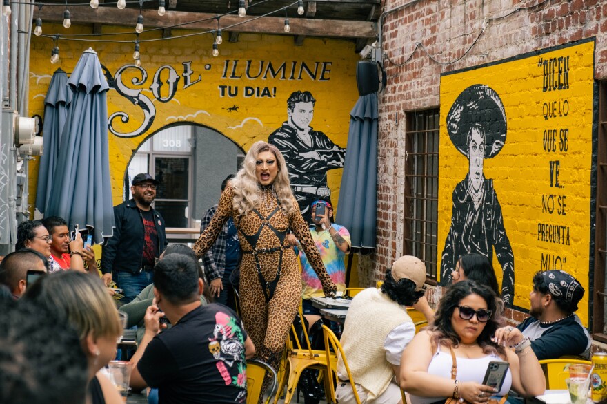 A feminine person wearing a cheetah print body suit and a blond wig walks through an outdoor patio with tables. A diverse group of people are eating and watching. The walls surrounding the outdoor patio are painted bright yellow and black and include an image of the singer Juan Gabriel and another man.