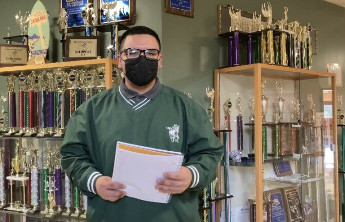 Young man with short dark hair and face mask stands in front of trophy case.