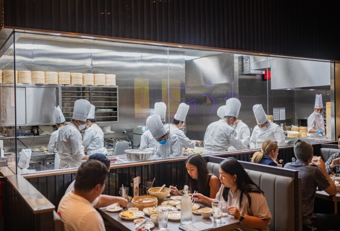 Men and women sit in booths while many chefs in white hats and jackets, behind a glass wall, are preparing food