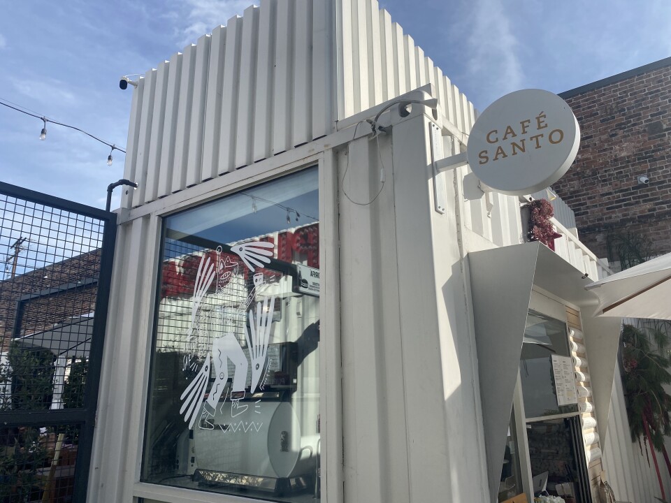 The exterior of a small coffee house is lined in a rustic, corrugated metal style: It's all painted light tan, and there's a circular sign that proclaims Café Santo in a reddish brown font. 