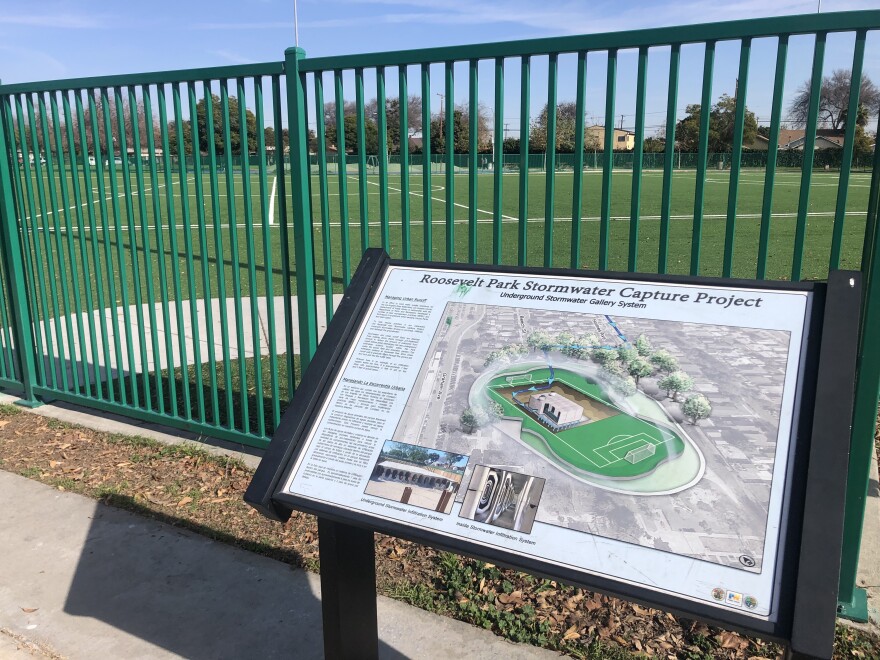 An educational sign in a park shows a diagram of a soccer field with a filtration system beneath it. In the background is a green fence surrounding a turf soccer field.