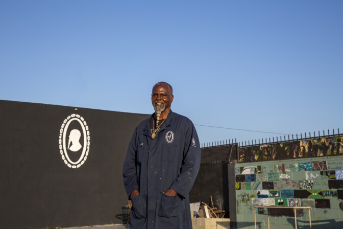 A Black man with a long white goatee wearing a long navy painter's coat, smiles at the camera with his hands in his front coat pockets. Behind him is a black wall with a white, curio-style portrait of him painted on it, and directly behind him is a cinder block wall with each block painted differently. Above him is a clear blue sky.