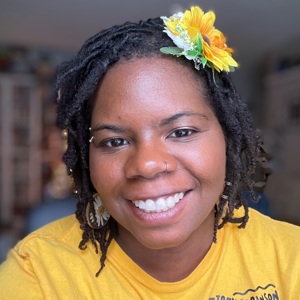 Antoinette Scully smiles at the camera. She is a Black woman with braided hair, dark eyes and a yellow flower in her hair. She is wearing a yellow shirt.