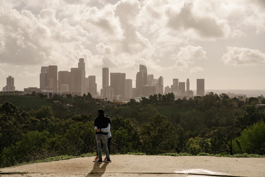 Two people embrace on the edge of a dirt road with the Los Angeles skyline in the background and dramatic clouds overhead mixed with sunlight, turning the sky a hazy yellowish gray.