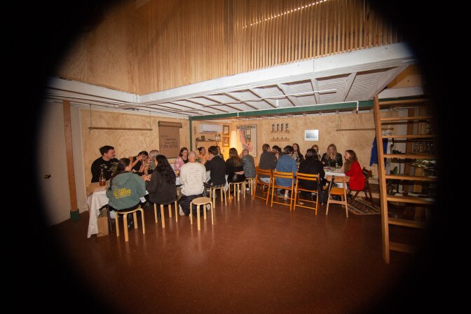 A peephole image of a group of people sitting on chairs and stools at an L-shape table under a loft area. The floor where they are seated is painted dark brown, and the loft walls are covered with light-brown wood paneling. There is a ladder positioned next to the group. 