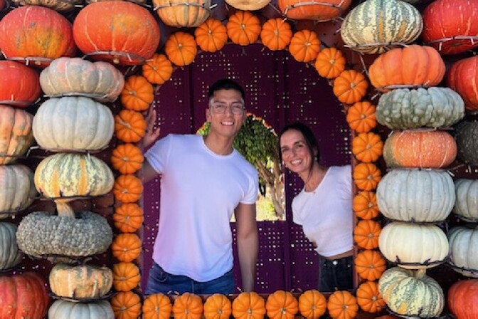 A white woman with brown hair and a Latino man in glasses in white t-shirts smile out from a little house covered in pumpkins 