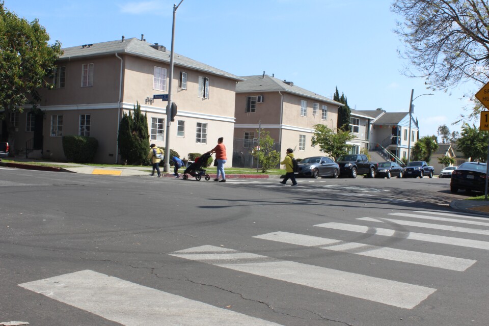 A woman pushes a stroller and children walk in front and behind her crossing a street.
