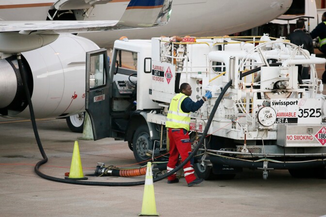 A man wearing a neon yellow safety vest and red pants stands beside a white work truck, parked beside an airplane.