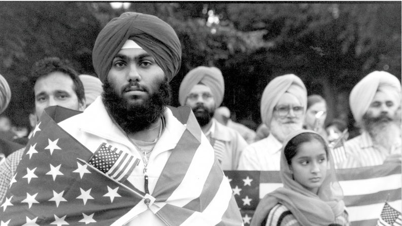 A Sikh man is wrapped in an American flag, while standing with a group at a vigil.