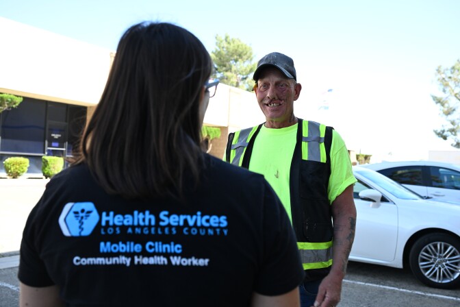 A person with dark hair wears a black shirt that says "Health Services, Los Angeles County, Mobile Clinic, Community Health Worker." She faces a man wearing a neon work uniform and gray hat. 