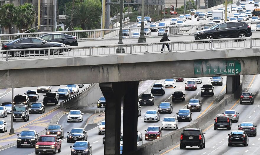 Cars crawl along the 110 Freeway through downtown Los Angeles as a person walks across on an overpass above.