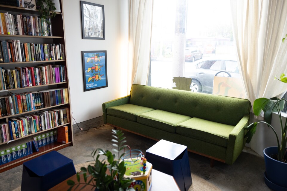 A brightly lit room with a big window behind a large green couch. In front of the couch there's a wooden table and dark blue stools. To the left of frame there's a large wooden bookcase full of books. 