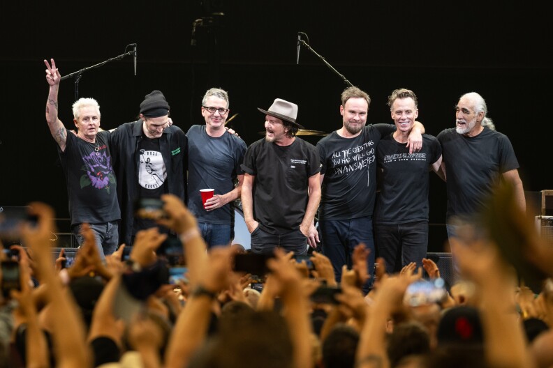 The members of the band Pearl Jam stand together in a row on stage with their arms around each other's shoulders while wearing black, casual clothing. They're taking in the audience cheering for them below.