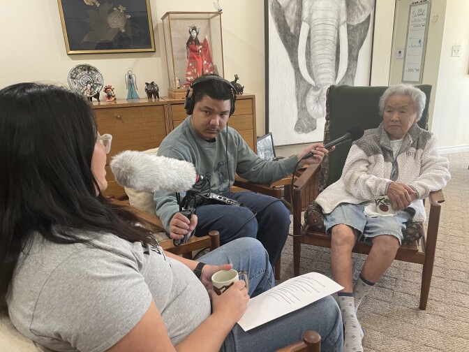 An older Asian woman with short white hair sits at a black chair, in the center an Asian man with short dark hair and green shirt holds a microphone towards the elder's face and another microphone to an Asian woman with dark hair and glasses that sits to the left of frame. 