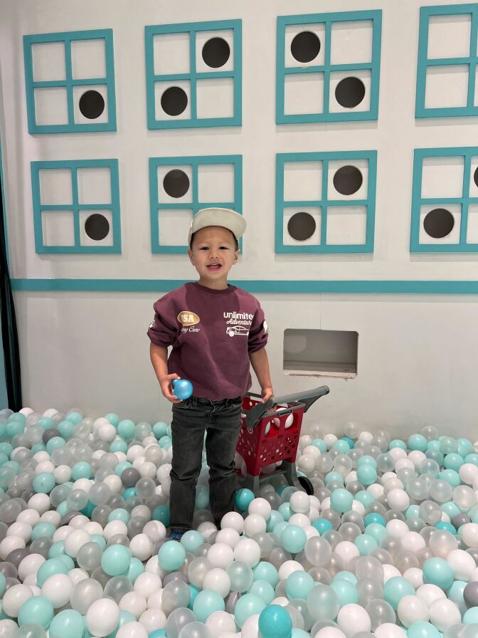 A young male wearing a maroon sweatshirt and dark pants holds a blue ball in his left hand and the handle of a toy shopping cart in the other hand. He is standing in a ball pit. 