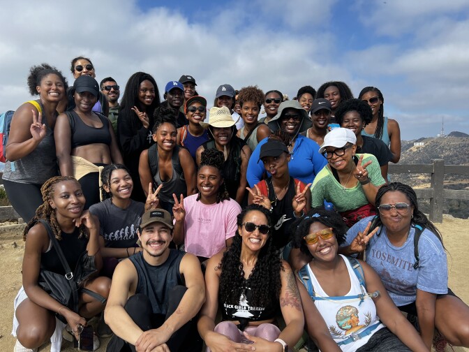 About two dozen hikers pose for a photo. Many of them are wearing hats or baseball caps to protect themselves from the sun. 