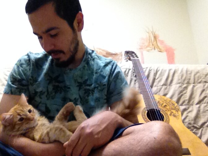 Frank Campos is seated holding a cat. A guitar sits to his left. 