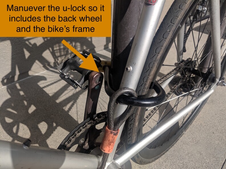 A close-up of a bike's tire with a bike lock on on the backside. There is an arrow pointing to it with text that reads, "manuever the u-lock so it includes the back wheel and the bike's frame."